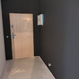 Tomasz Adamski - Świeżo pomalowane na grafitowo ściany w korytarzu, zabezpieczona folią lampa, drzwi wejściowe z widocznymi śladami po remoncie, podłoga w trakcie prac wykończeniowych.