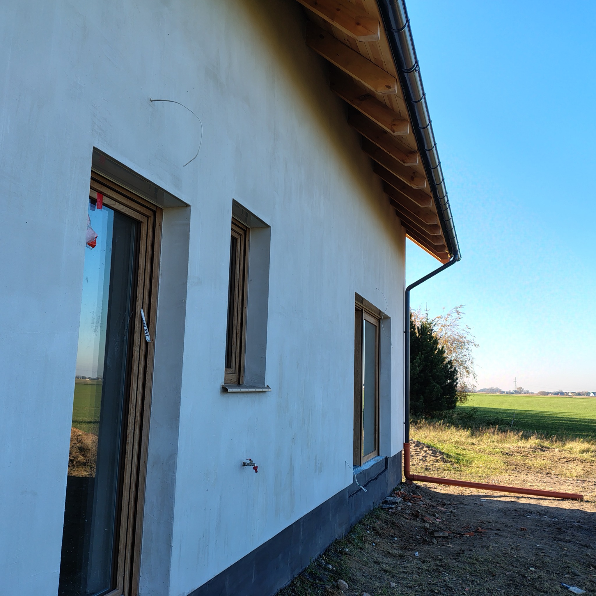 Elewacja domu jednorodzinnego po tynkowaniu, z widocznymi oknami z drewnianymi ramami, rynną i fragmentem krajobrazu z polami w tle. Świeżo otynkowana ściana.