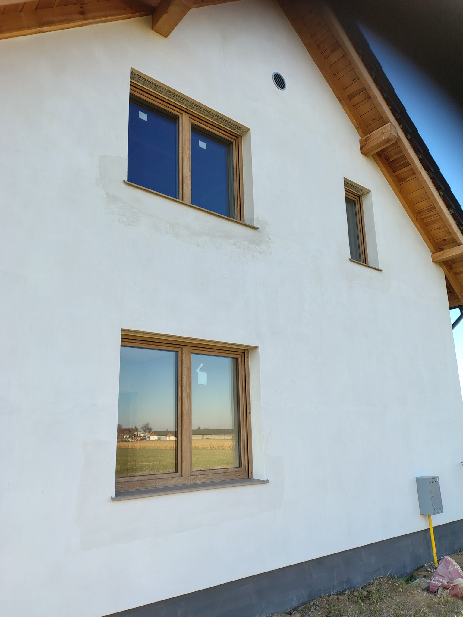 Biała elewacja domu z drewnianymi oknami, widoczne ślady po łataniu tynku. W oknach odbija się krajobraz. Szara podmurówka i wystające elementy dachu z drewna.