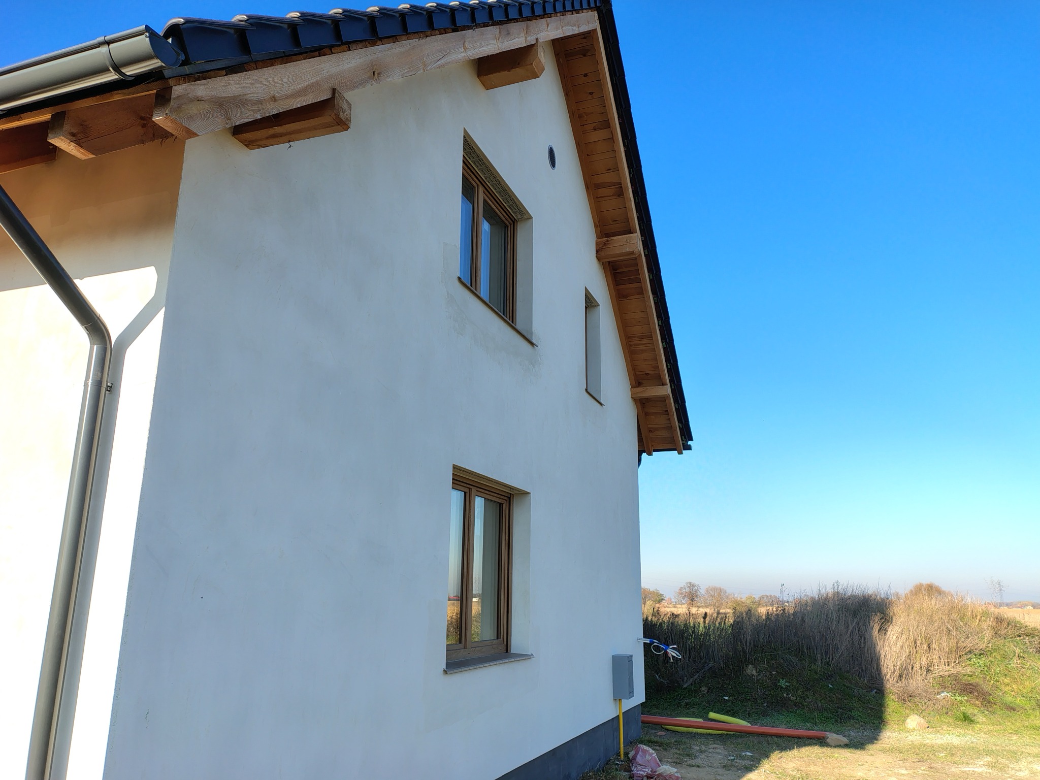 Boczny widok otynkowanego domu jednorodzinnego z drewnianymi elementami wykończeniowymi dachu i brązowymi oknami na tle błękitnego nieba.