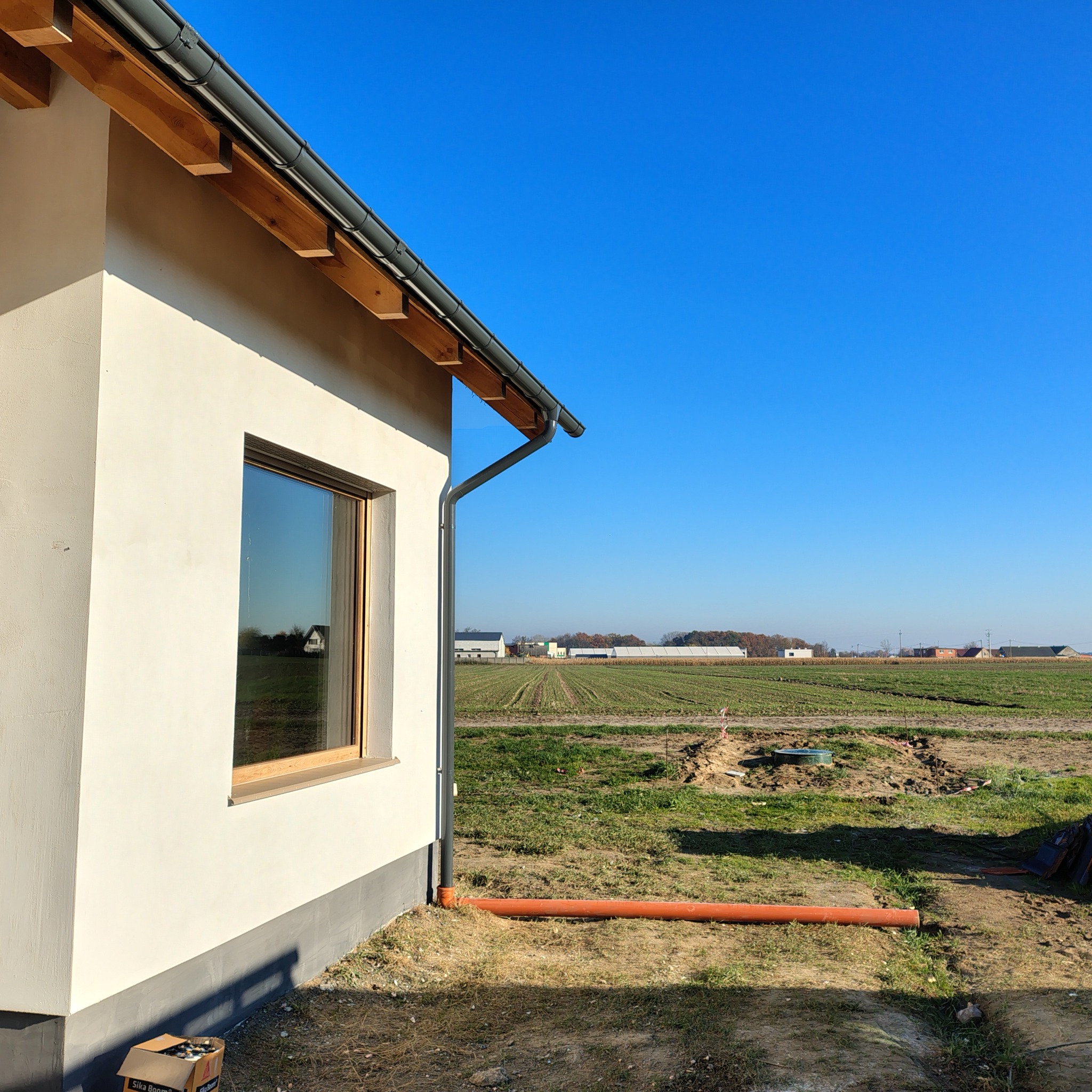 Elewacja domu jednorodzinnego z oknem i orynnowaniem, widok z zewnątrz na pole uprawne. W tle zabudowania gospodarcze. Pomarańczowa rura drenażowa przy ścianie.