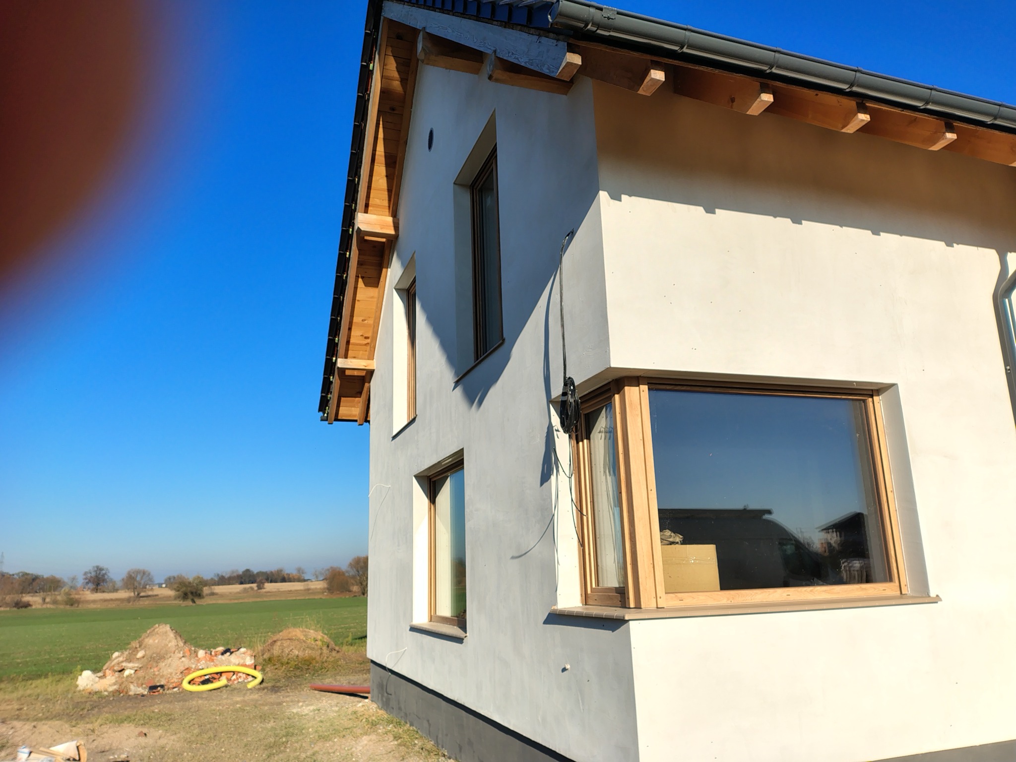 Elewacja domu jednorodzinnego w trakcie prac wykończeniowych, widoczne okna z drewnianą stolarką i fragment elewacji w szarym kolorze. W tle zielone pole i błękitne niebo.