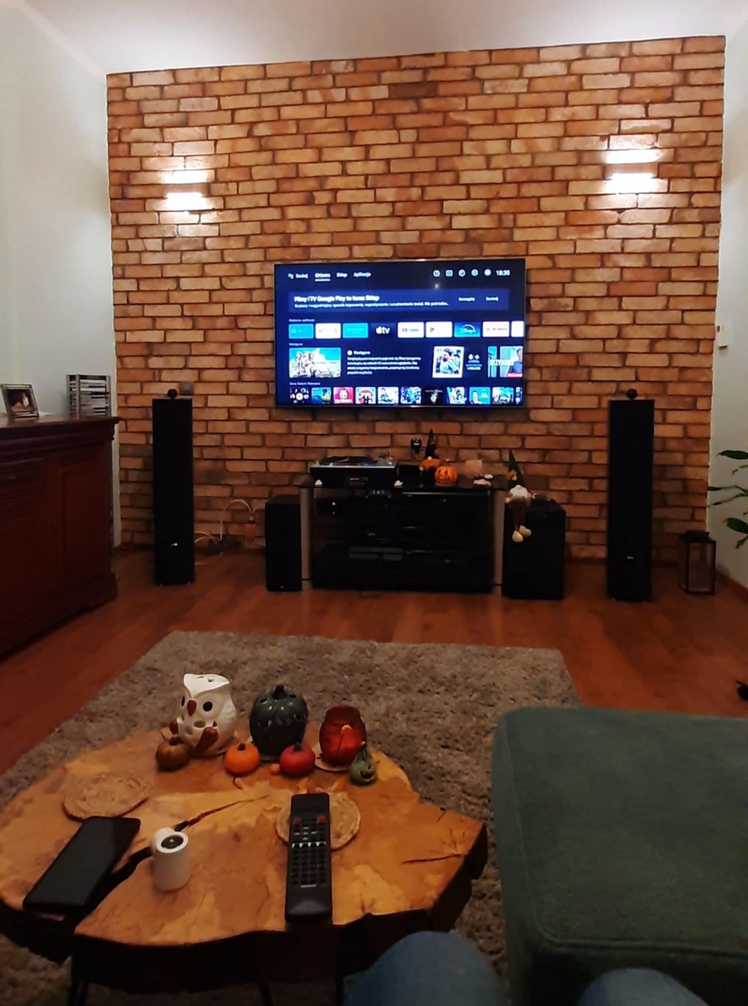 Ściana z cegły klinkierowej za telewizorem, oświetlona kinkietami. Element wykończenia wnętrza salonu z drewnianą podłogą i stolikiem kawowym.