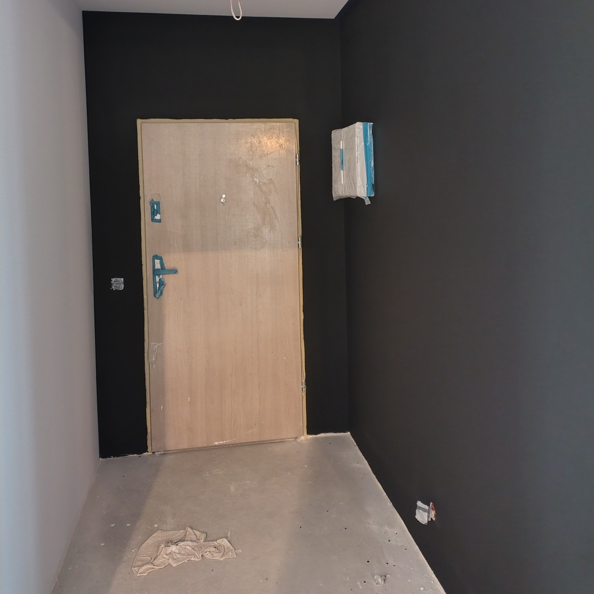 Świeżo pomalowane na czarno ściany w pokoju z surową podłogą i zabezpieczonymi elementami. Widoczna drewniana ościeżnica i drzwi. Etap prac wykończeniowych.