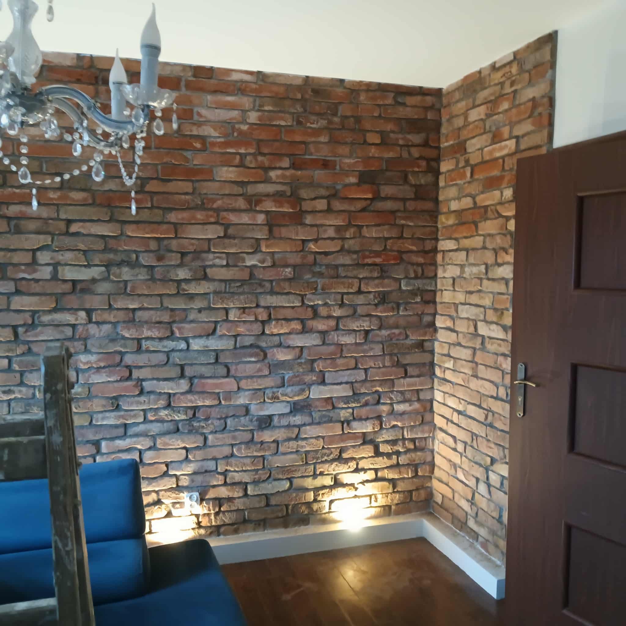 Ściana z cegły dekoracyjnej z oświetleniem punktowym u dołu, widoczny fragment żyrandola i drzwi. Element wykończenia wnętrza w stylu industrialnym.