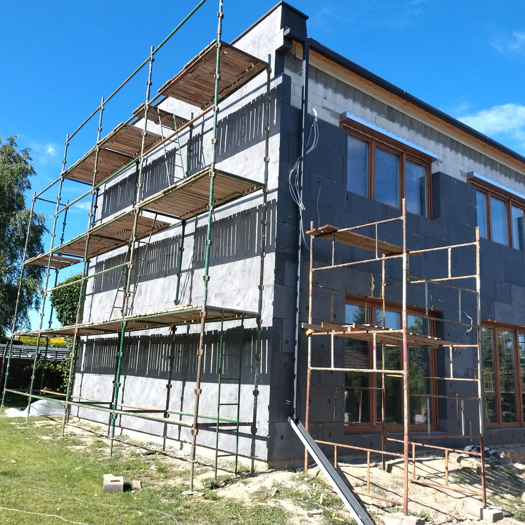 Budynek w trakcie ocieplania styropianem, widoczne rusztowania i okna. Część elewacji szara, część czarna. Słoneczny dzień, błękitne niebo.