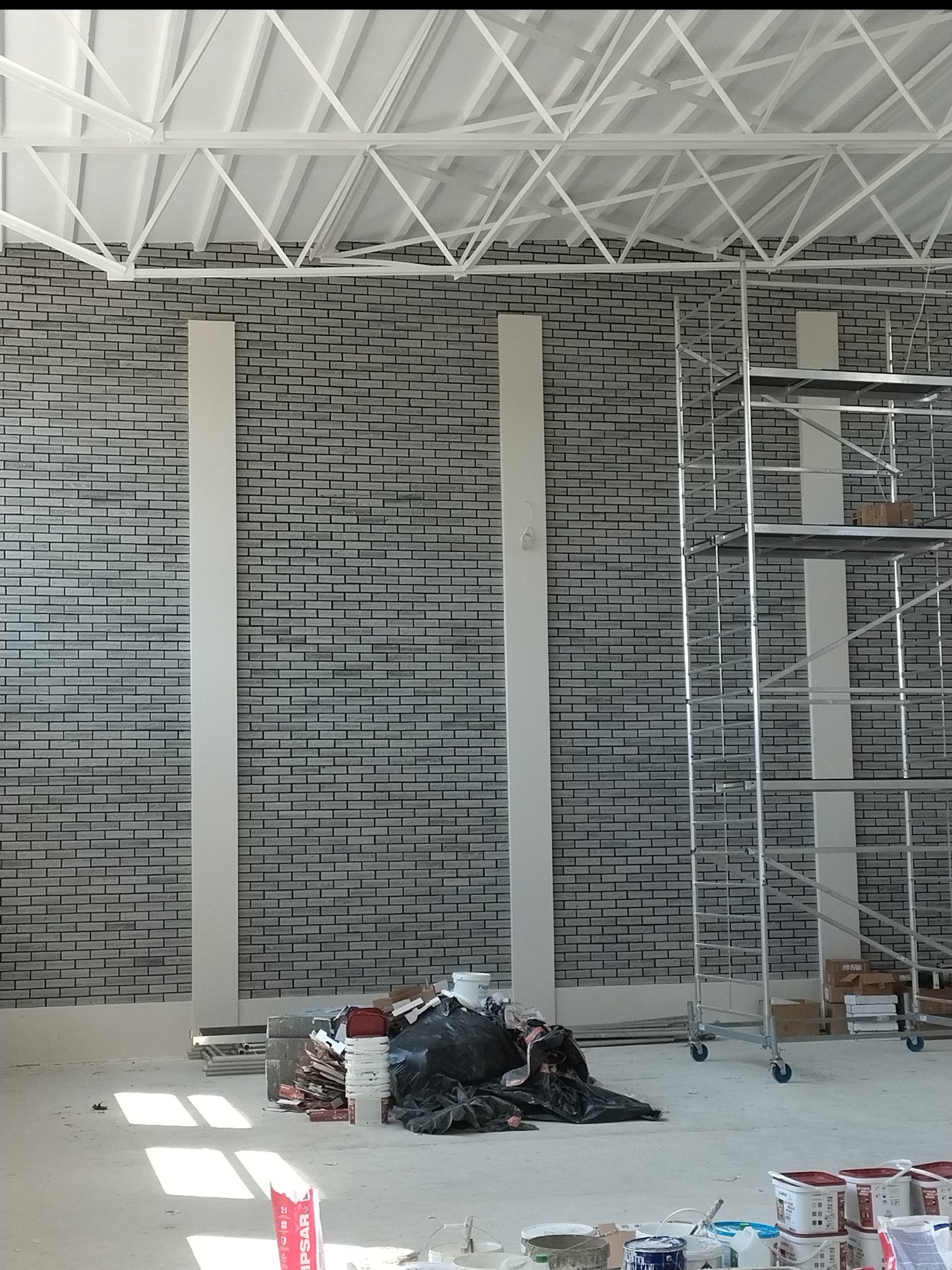 Wnętrze w trakcie prac wykończeniowych: szara ceglana ściana z białymi pilastrami, rusztowanie i materiały budowlane na podłodze. Widoczna konstrukcja dachu.