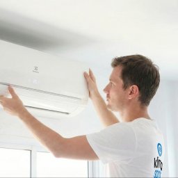 DM GROUP POLAND SPÓŁKA Z OGRANICZONĄ ODPOWIEDZIALNOŚCIĄ - Mężczyzna instaluje białą klimatyzację Electrolux w jasnym pomieszczeniu. Widoczny wyświetlacz z temperaturą 20 stopni. Na koszulce logo firmy.