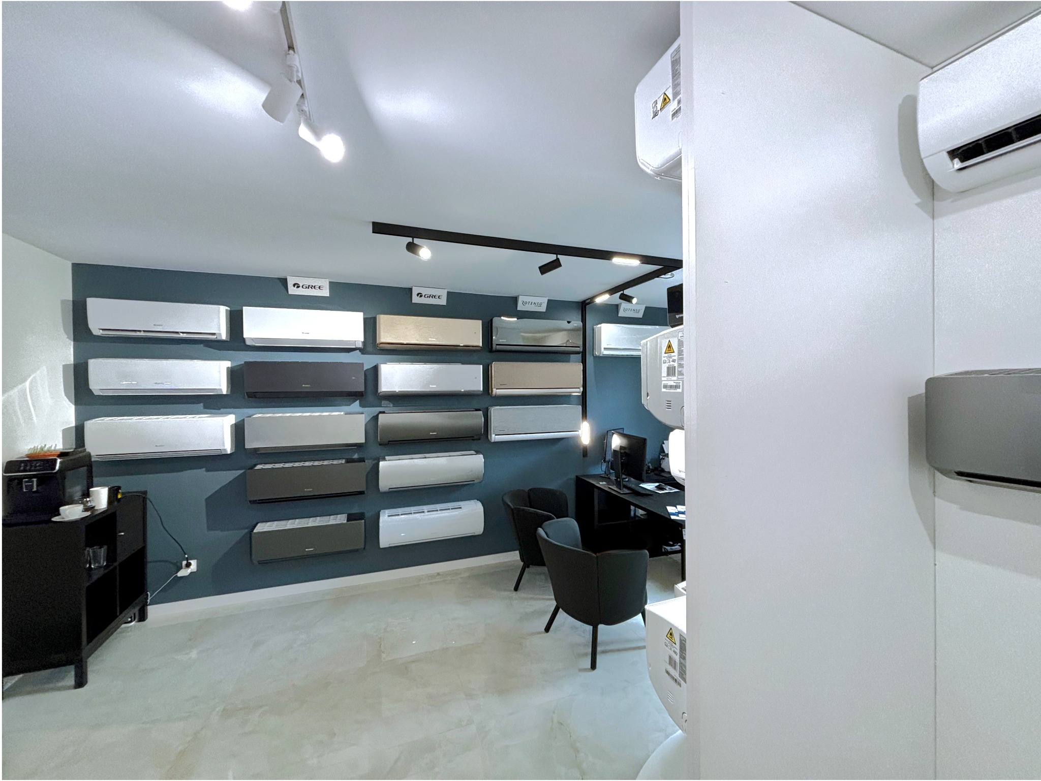 Ekspozycja klimatyzatorów różnych marek i kolorów na ścianie w salonie. Widoczne modele Gree i Rotenso, oświetlenie punktowe i biurko z komputerem.