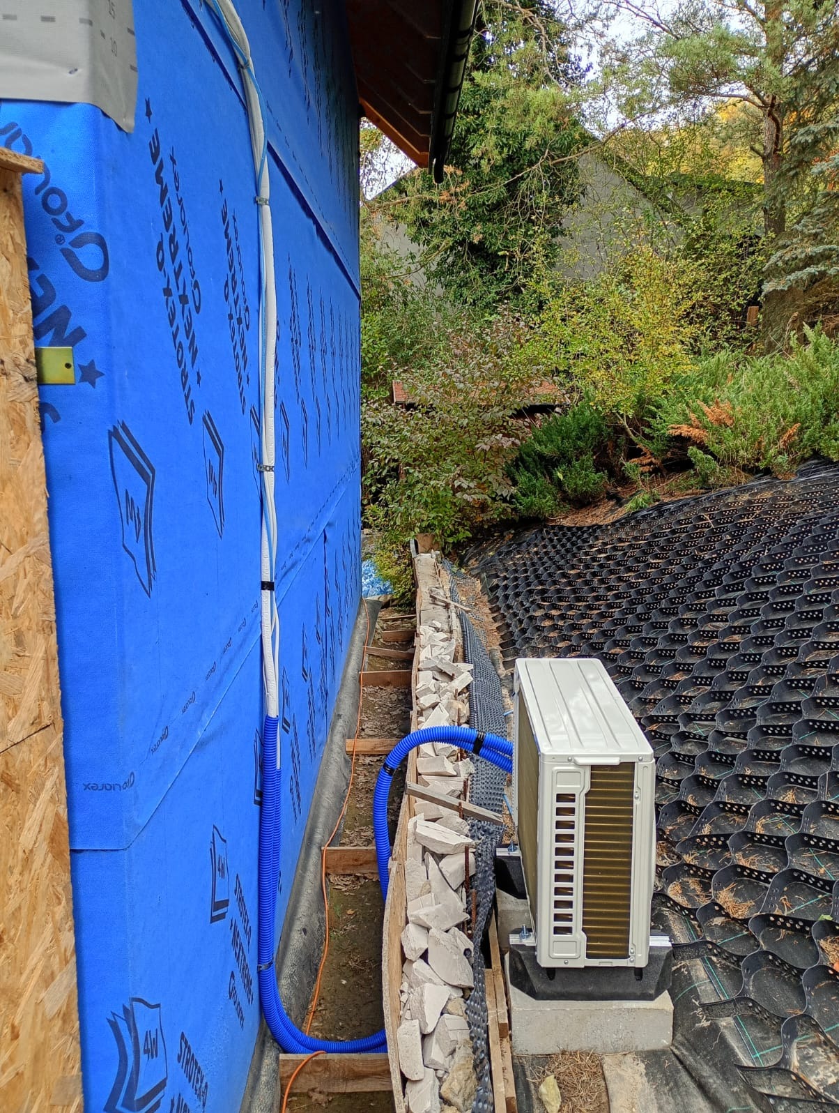 Montaż zewnętrznej jednostki klimatyzacji na dachu z niebieską izolacją ściany. Widoczne białe i niebieskie rury instalacyjne. Otoczenie: zieleń, elementy konstrukcyjne.