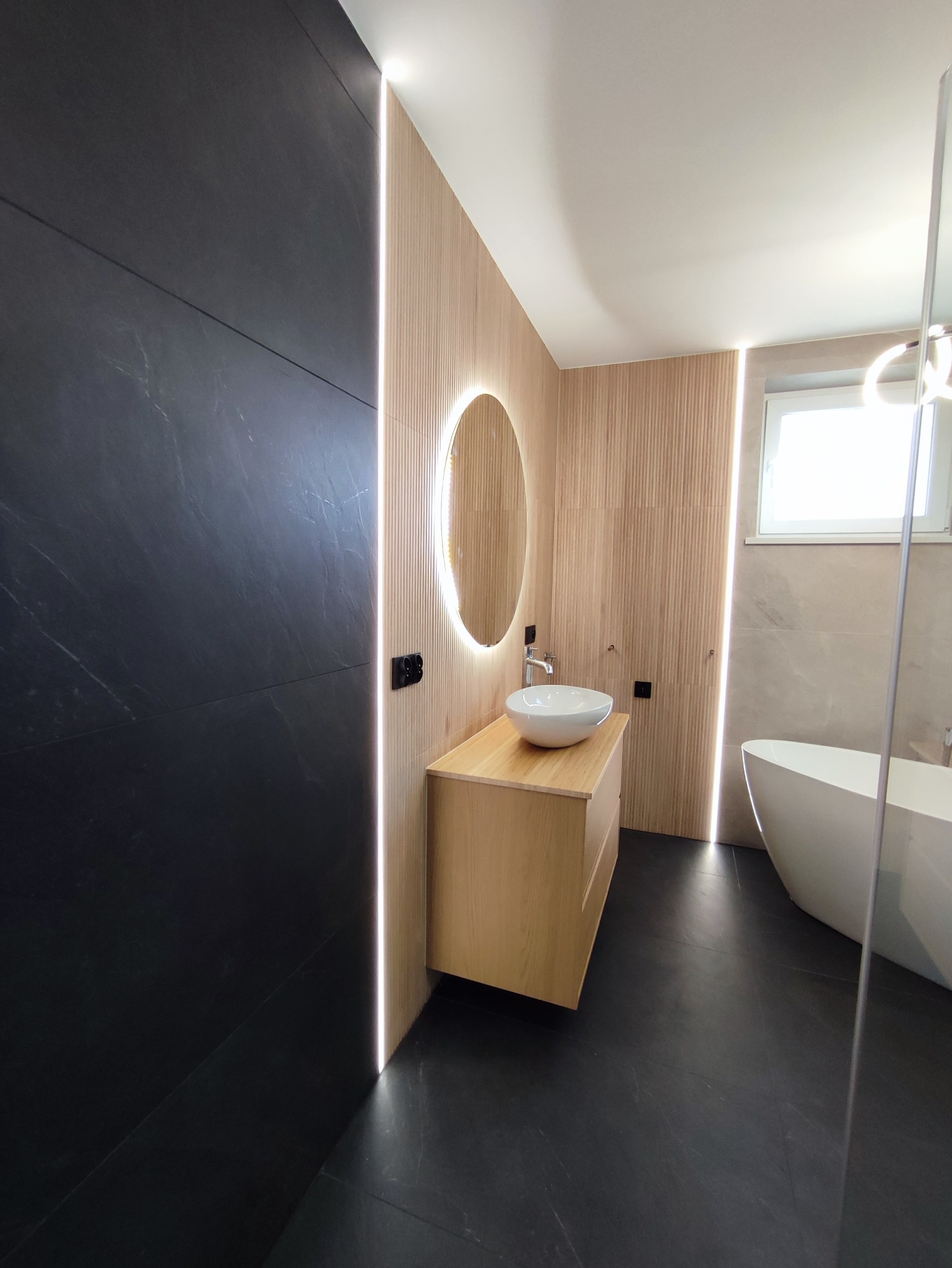 Nowoczesna łazienka z czarnymi płytkami, drewnianą zabudową umywalki i podświetlanym lustrem. Minimalistyczny design z akcentami świetlnymi.