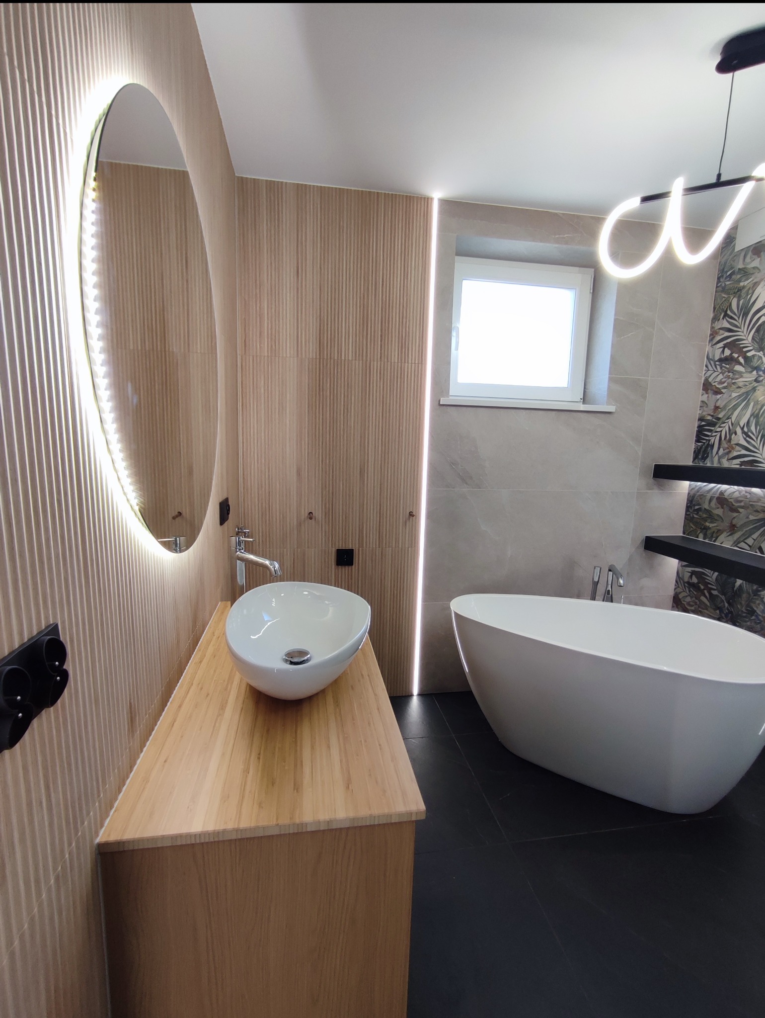 Nowoczesna łazienka z owalną umywalką na drewnianej szafce, okrągłym lustrem z podświetleniem LED i wolnostojącą wanną. Minimalistyczny design z akcentami drewna i roślin.