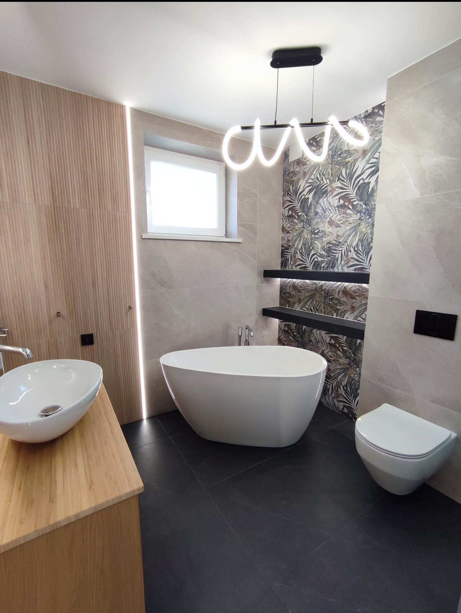 Nowoczesna łazienka z wolnostojącą wanną, umywalką na drewnianej szafce i toaletą. Na ścianie płytki z motywem roślinnym i podświetlane półki. Całość oświetla designerska lampa.