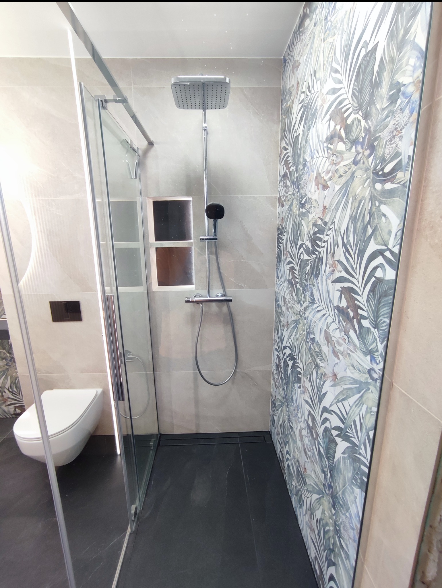 Nowoczesna kabina prysznicowa typu walk-in z deszczownicą, wykończona płytkami w odcieniach szarości i beżu, z akcentem dekoracyjnej ściany w motywy roślinne.