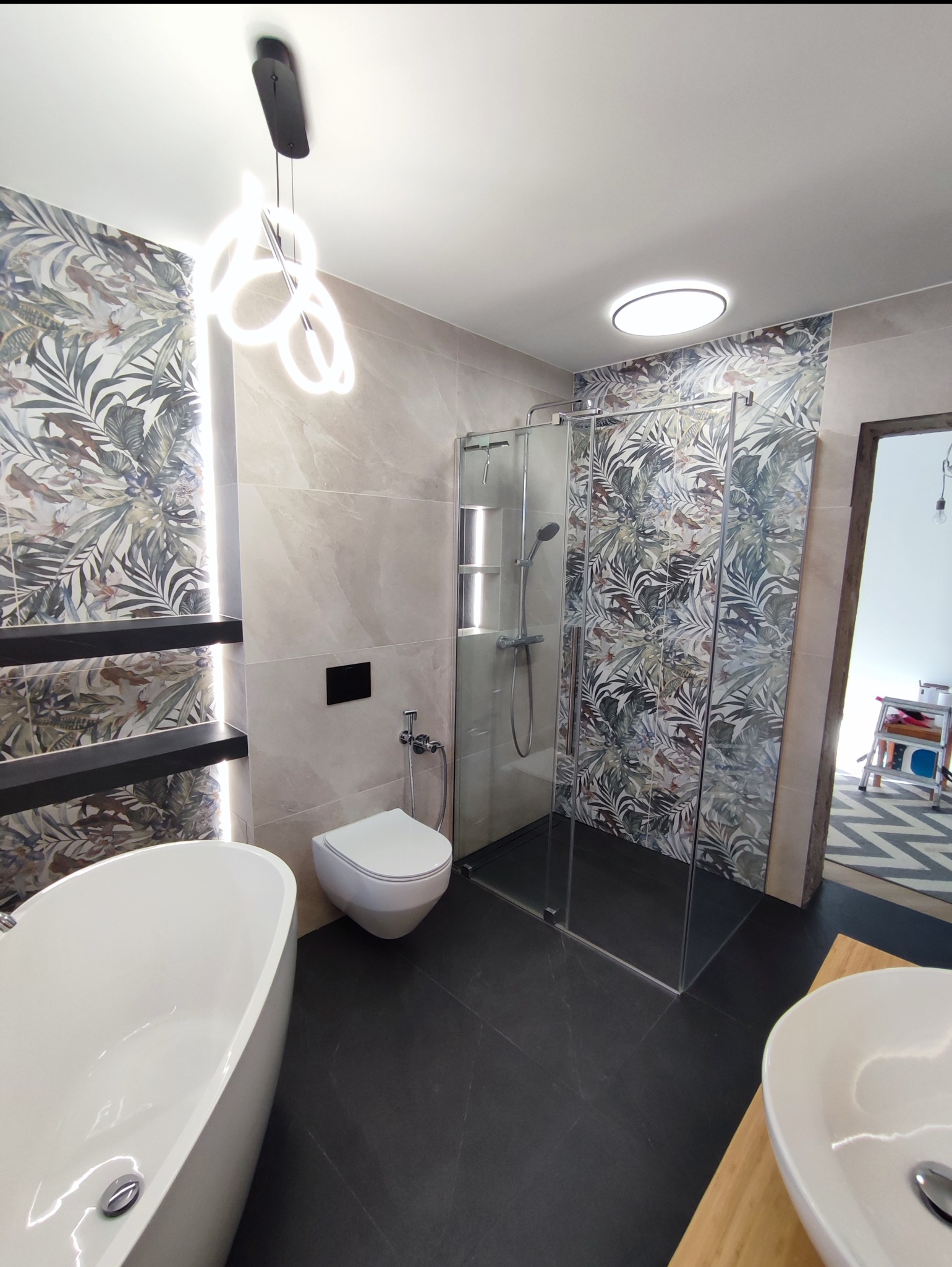 Nowoczesna łazienka z wanną wolnostojącą, kabiną prysznicową i toaletą. Na ścianach płytki z motywem roślinnym i marmurkowe. Oświetlenie LED. Czarna podłoga.