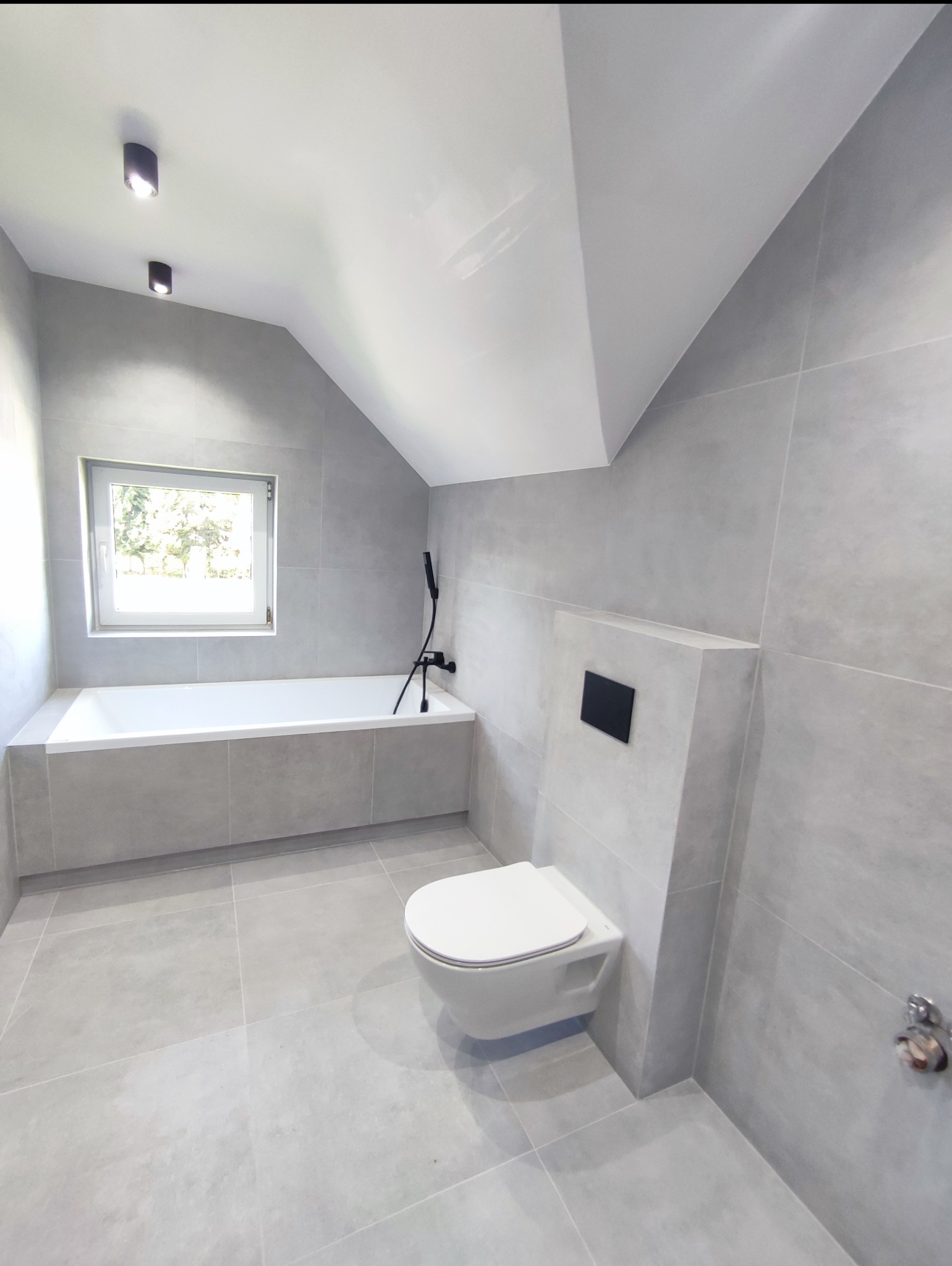Jasna łazienka na poddaszu w szarościach: wanna pod oknem, podwieszana toaleta z czarnym przyciskiem, płytki wielkoformatowe, minimalistyczne oświetlenie.