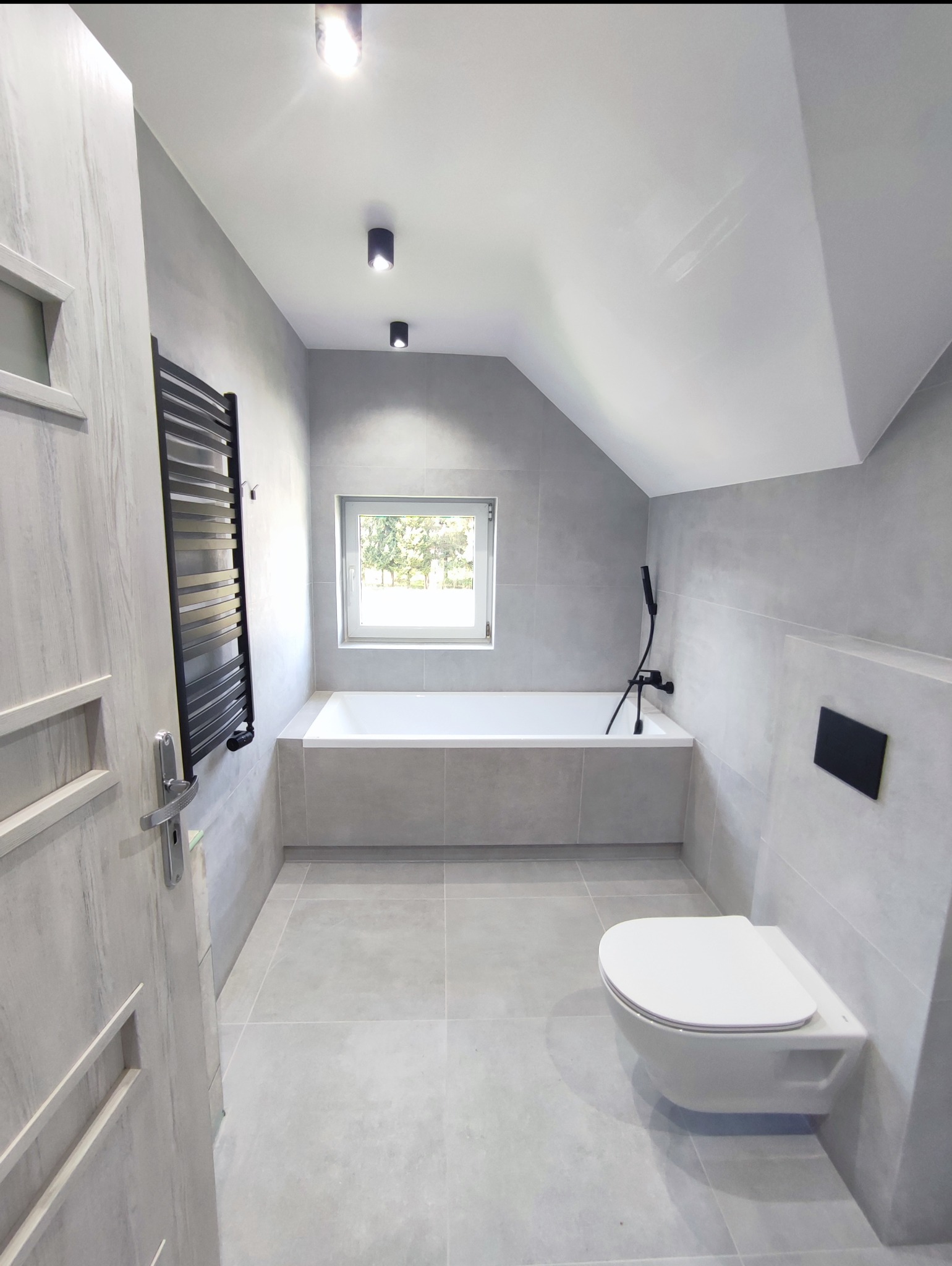 Jasna łazienka na poddaszu z wanną, toaletą podwieszaną i grzejnikiem drabinkowym. Szare płytki na ścianach i podłodze. Widok przez okno na zieleń.