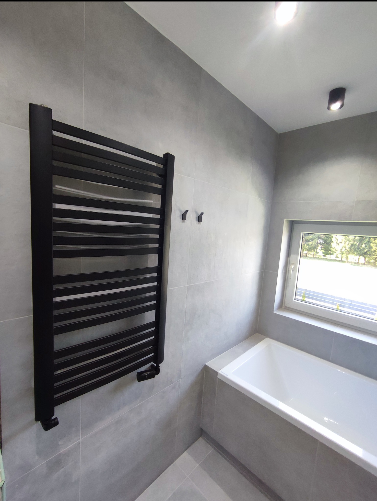 Nowoczesna łazienka w szarościach z czarnym grzejnikiem drabinkowym, widok na wannę i okno. Minimalistyczny design, płytki wielkoformatowe, oświetlenie punktowe.