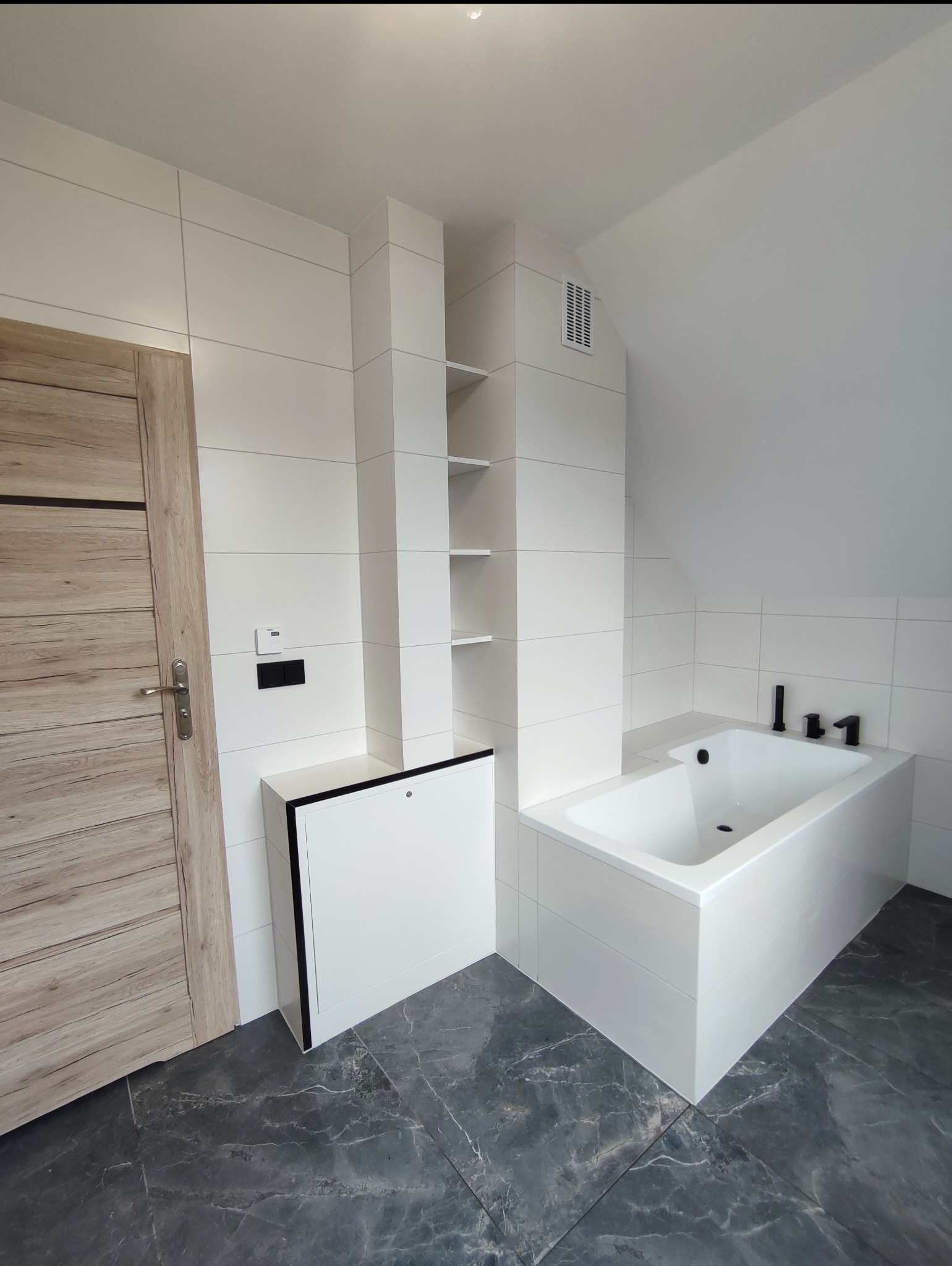 Nowoczesna łazienka w Górkach Wielkich po remoncie. Białe płytki, czarna armatura, szara podłoga z marmurkowym wzorem. Minimalistyczny design, zabudowa wnęki z półkami.