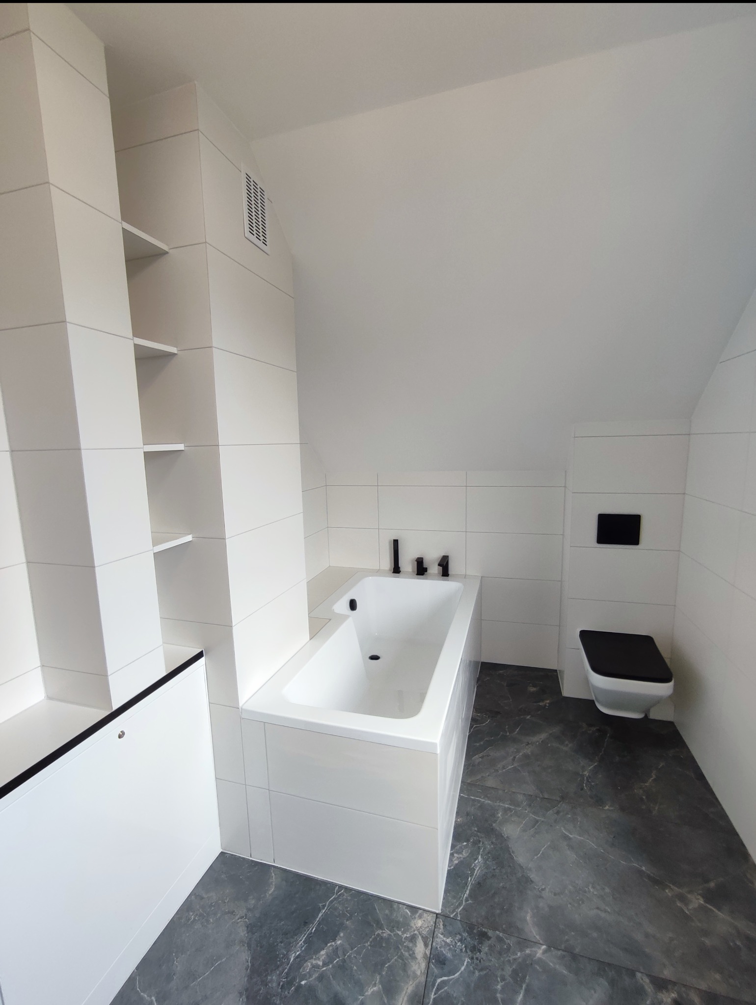 Nowoczesna łazienka na poddaszu z białą wanną, toaletą w kolorze czerni i bieli oraz szarą marmurową podłogą. Ściany wyłożone białymi, prostokątnymi płytkami.