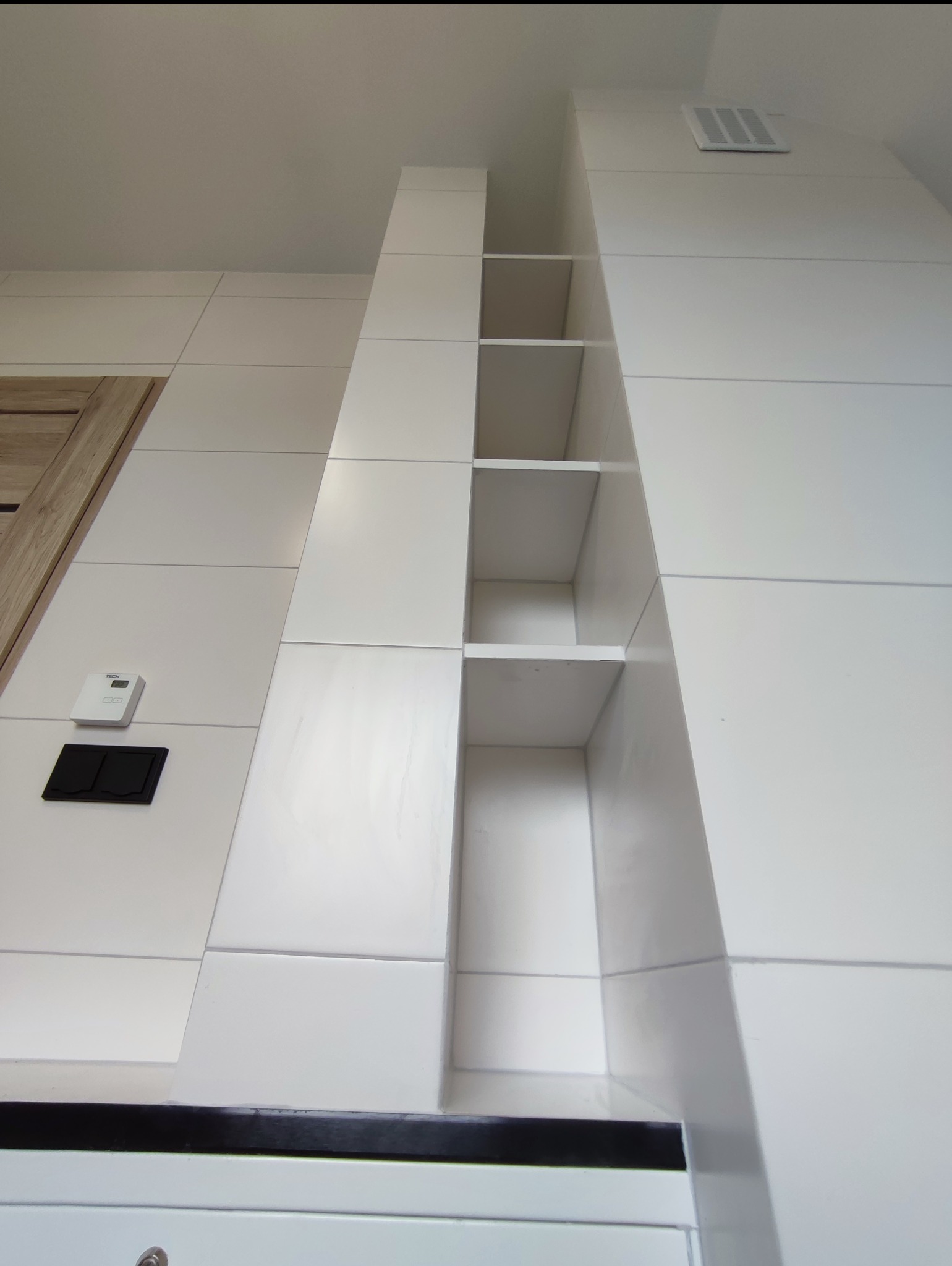 Biała, nowoczesna łazienka z półkami wnękowymi, wykończona płytkami w dużym formacie. Widoczny fragment drzwi i panel sterowania. Ujęcie z dołu.