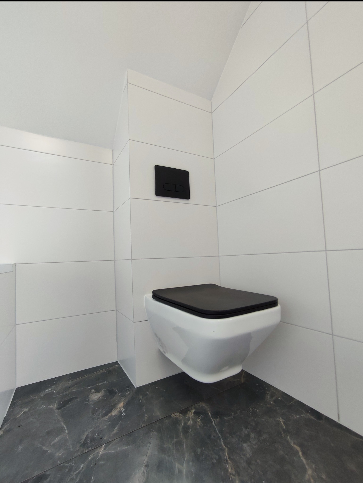 Nowoczesna łazienka z białymi płytkami i czarną armaturą. Podwieszana toaleta z czarną deską na tle marmurowej podłogi. Minimalistyczny design.