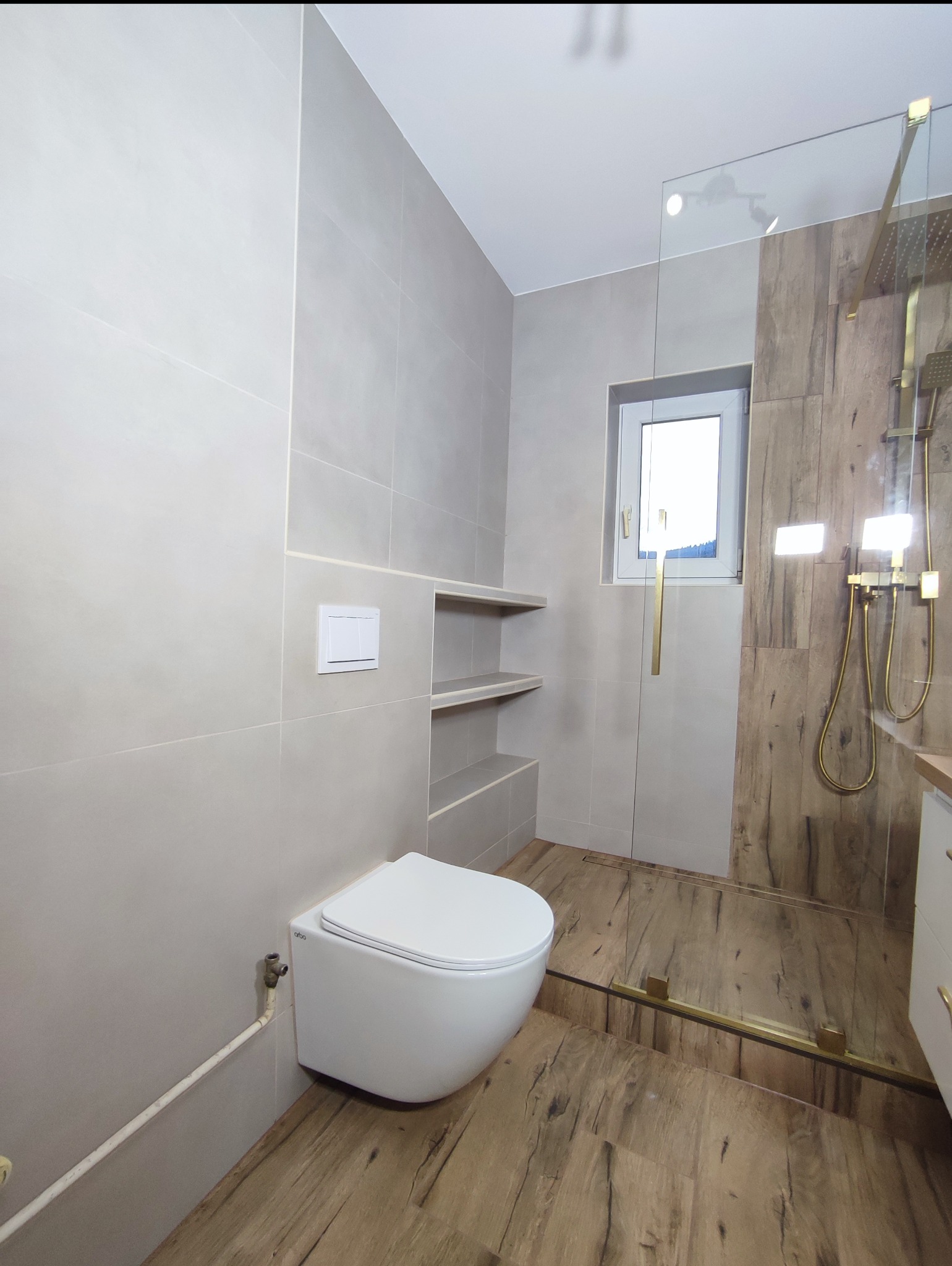 Nowoczesna łazienka z beżowymi płytkami, drewnopodobną podłogą i złotymi akcentami. Kabina prysznicowa ze szklaną ścianką i deszczownicą. Wisząca toaleta i wnęki w ścianie.