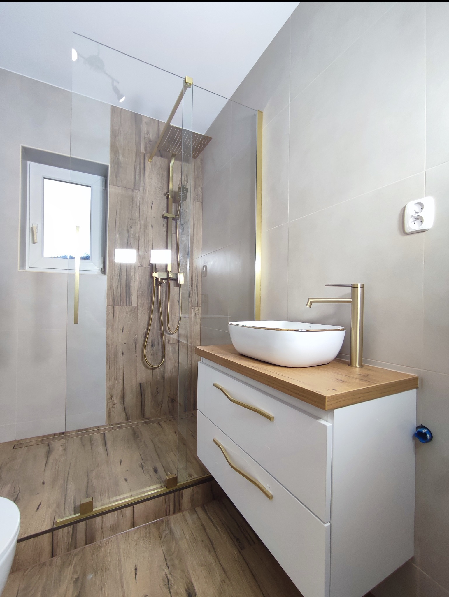 Nowoczesna łazienka z kabiną prysznicową typu walk-in, umywalką nablatową na szafce z drewnianym blatem i złotą armaturą. Jasne płytki i drewnopodobna podłoga.