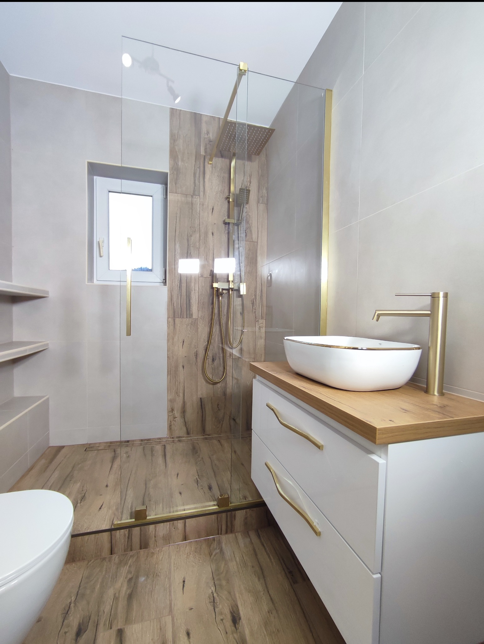 Nowoczesna łazienka z kabiną prysznicową typu walk-in, drewnopodobnymi płytkami i złotymi akcentami. Biała umywalka nablatowa na szafce z drewnianym blatem.