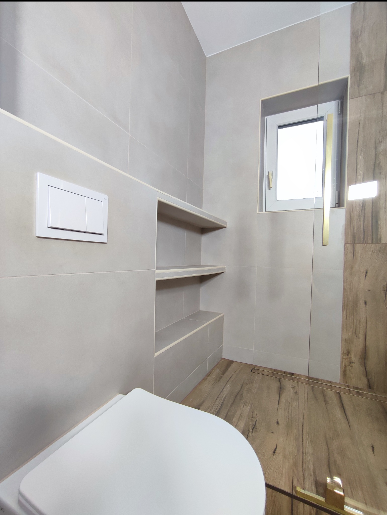 Nowoczesna łazienka z szarymi płytkami, drewnopodobną podłogą i wnękami na ścianie. Widoczny biały sedes i okno z jasnym światłem dziennym.