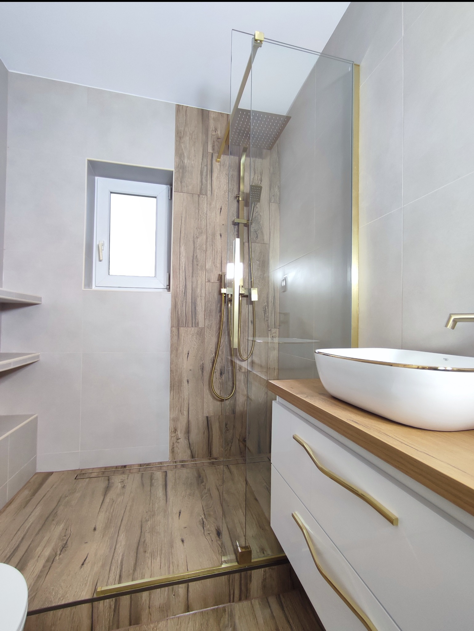 Nowoczesna łazienka z kabiną prysznicową typu walk-in, drewnopodobnymi płytkami i złotą armaturą. Umywalka nablatowa na szafce z białymi frontami i złotymi uchwytami.