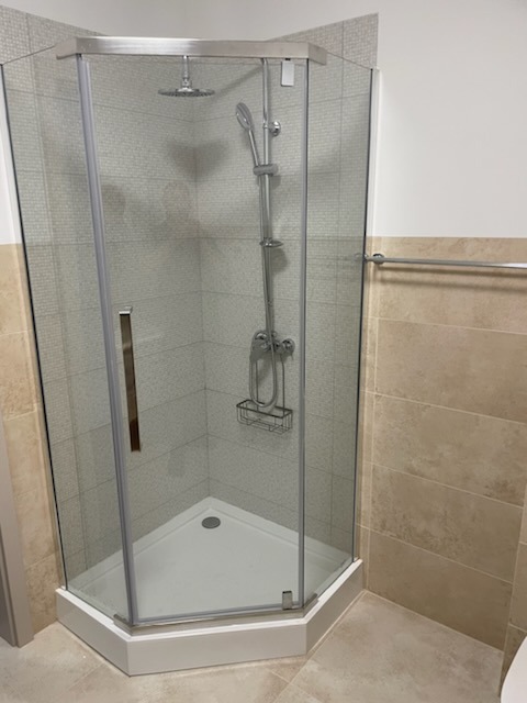 Narożna kabina prysznicowa z chromowanymi elementami i deszczownicą, wykończona beżowymi płytkami. Widoczny odpływ i półka na kosmetyki.