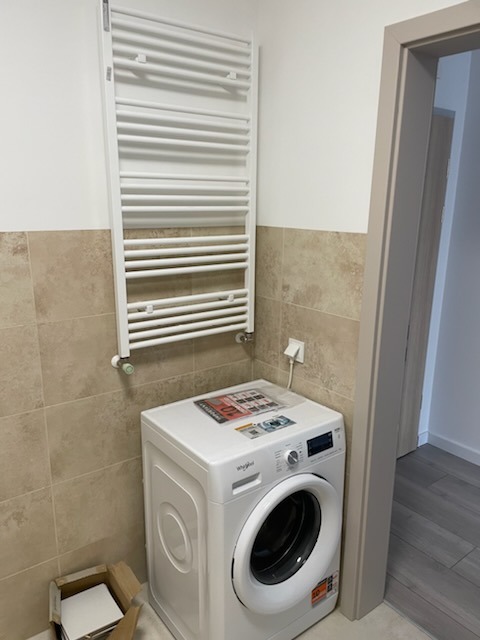 Biała pralka Whirlpool w łazience z beżowymi płytkami, obok grzejnik drabinkowy i otwarte drzwi. Widoczny karton z płytkami. Ujęcie z bliska, neutralne światło.