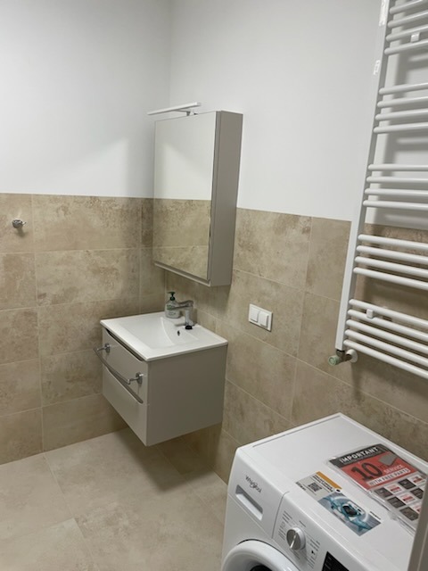 Jasna łazienka po remoncie: beżowe płytki, podwieszana szafka z umywalką, lustro z oświetleniem, grzejnik drabinkowy i pralka. Minimalistyczny design.