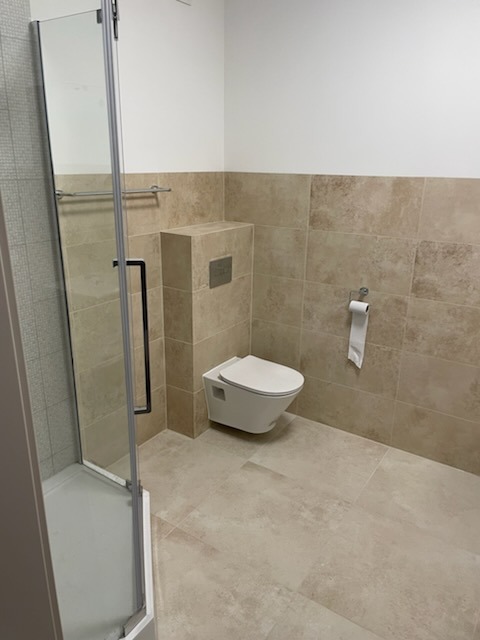 Nowoczesna łazienka z beżowymi płytkami, wiszącą toaletą i kabiną prysznicową z czarnym uchwytem. Minimalistyczny design, jasne wnętrze.