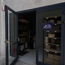 TALERON SPÓŁKA Z OGRANICZONĄ ODPOWIEDZIALNOŚCIĄ - Ciemne, częściowo otwarte podwójne drzwi do obiektu logistycznego, ukazujące wnętrze z regałami pełnymi kartonów i widocznym logo firmy spedycyjnej.