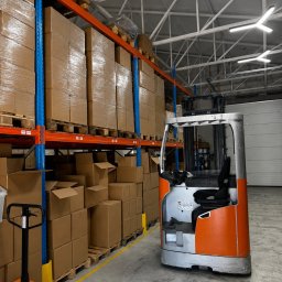 TALERON SPÓŁKA Z OGRANICZONĄ ODPOWIEDZIALNOŚCIĄ - Wysokie regały magazynowe wypełnione kartonowymi pudłami, częściowo owiniętymi folią, obok pomarańczowo-srebrny wózek widłowy typu reach truck i czarny wózek paletowy PROMAG.