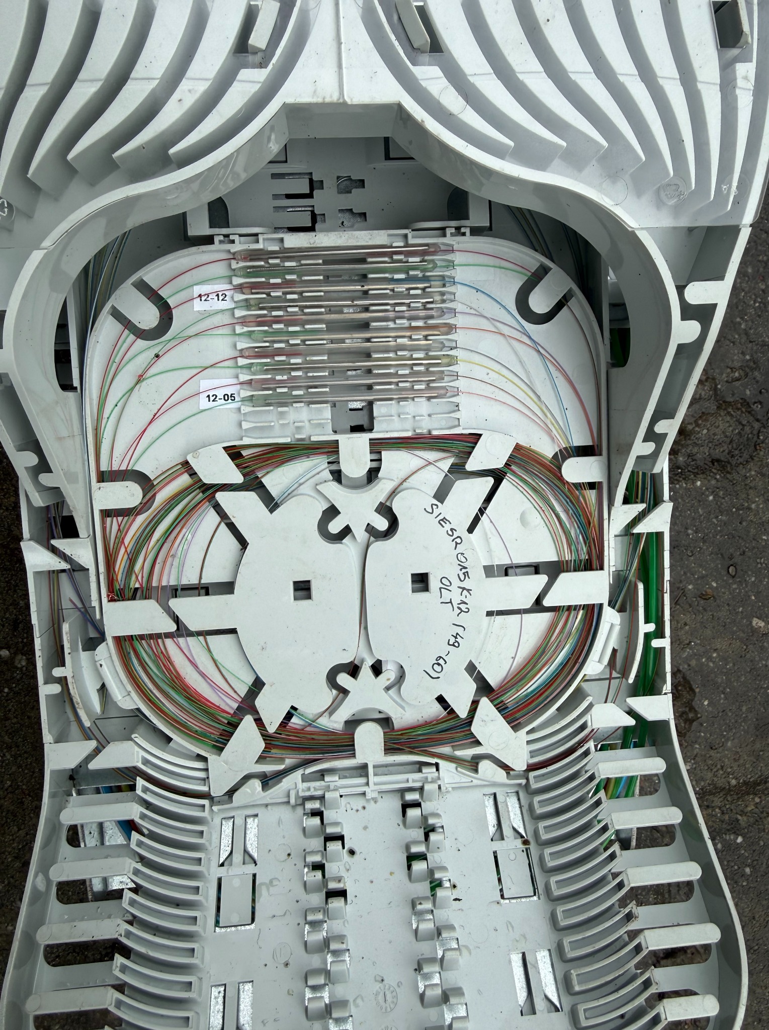 Otwarta, biała skrzynka telekomunikacyjna z precyzyjnie ułożonymi, kolorowymi kablami światłowodowymi. Widoczne oznaczenia na kablach i elementy mocujące.