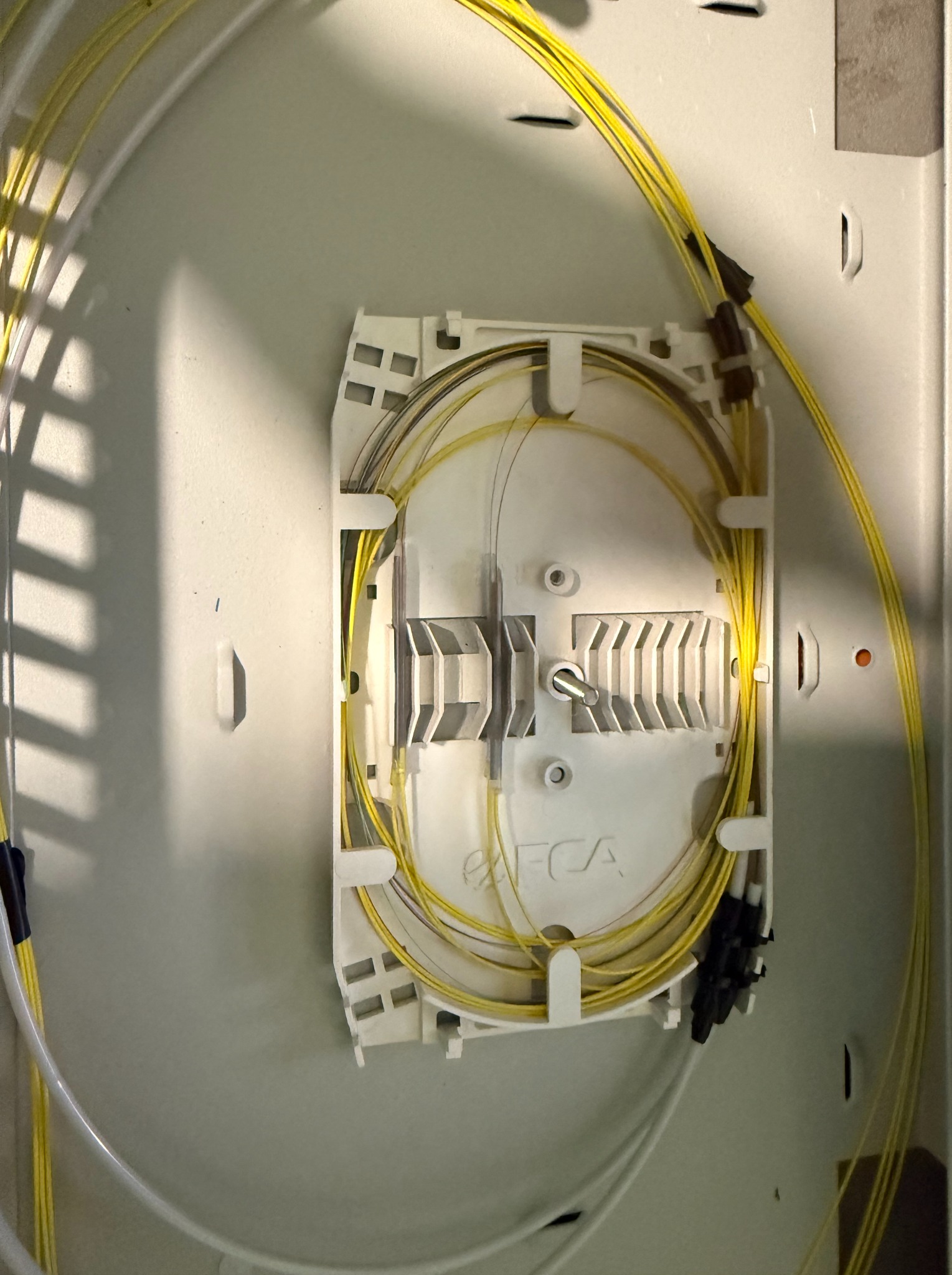 Biała skrzynka z organizerem światłowodów i żółtymi kablami światłowodowymi. Widoczne złącza i mocowania kabli. Całość w szarej obudowie.