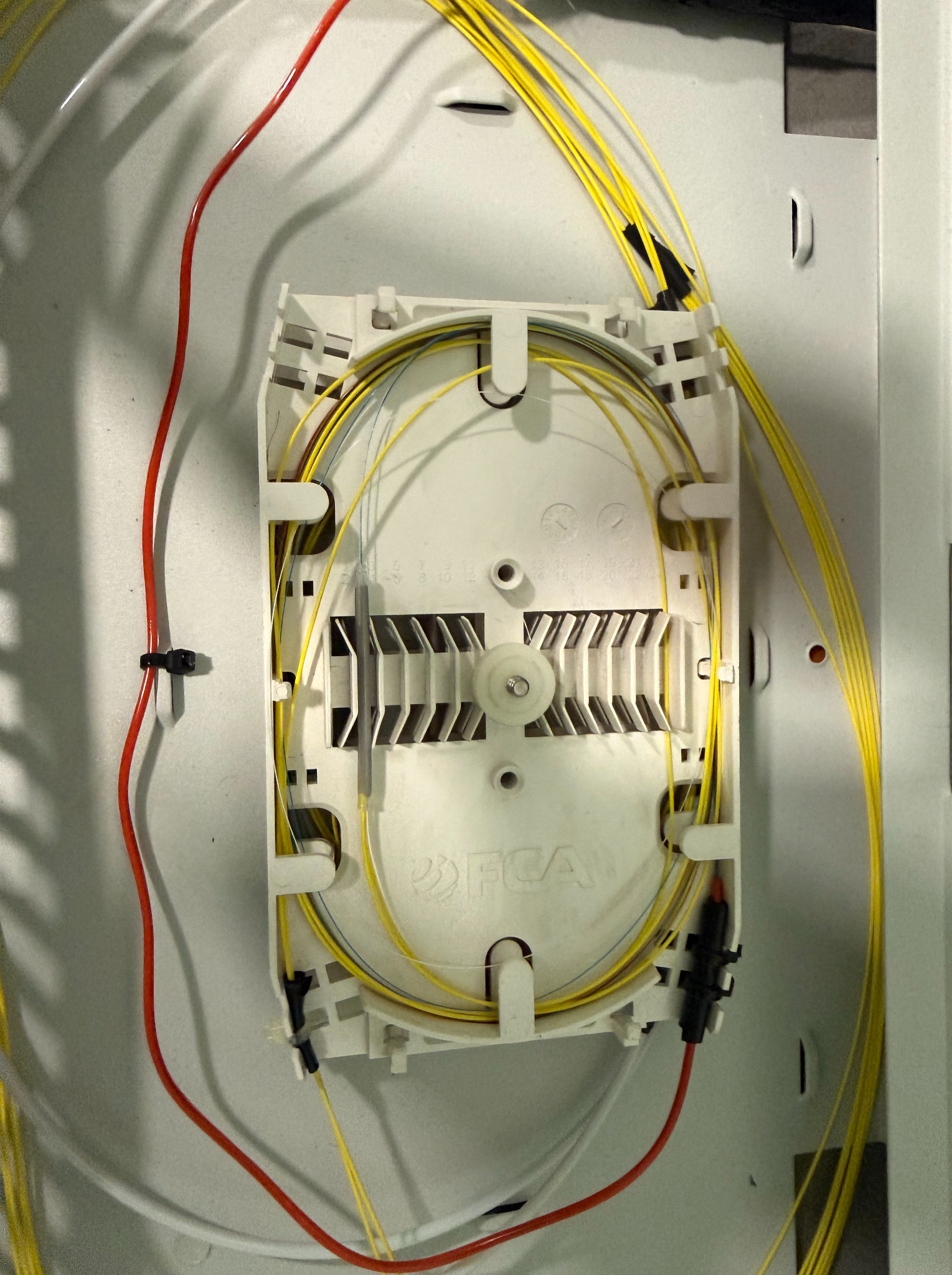 Rozdzielnica światłowodowa w białej obudowie z widocznymi kablami światłowodowymi w kolorze żółtym i czerwonym. Widoczne złącza i elementy mocujące.