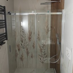 Niko Instalbud - Nowoczesna kabina prysznicowa z motywem roślinnym na szklanych drzwiach, w jasnej łazience z marmurowymi płytkami i drewnianymi panelami dekoracyjnymi.