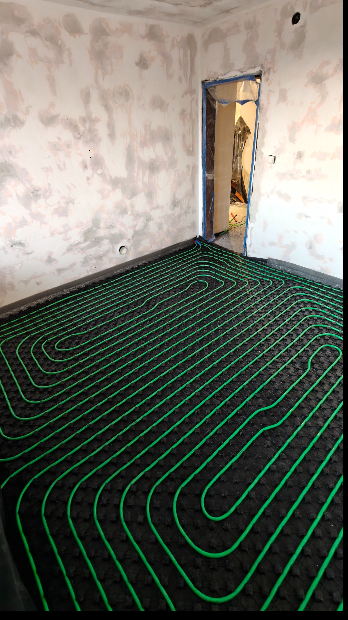 Zielone rury ogrzewania podłogowego ułożone na czarnej macie w surowym wnętrzu z przygotowanymi ścianami do malowania. Widok na próg drzwi.