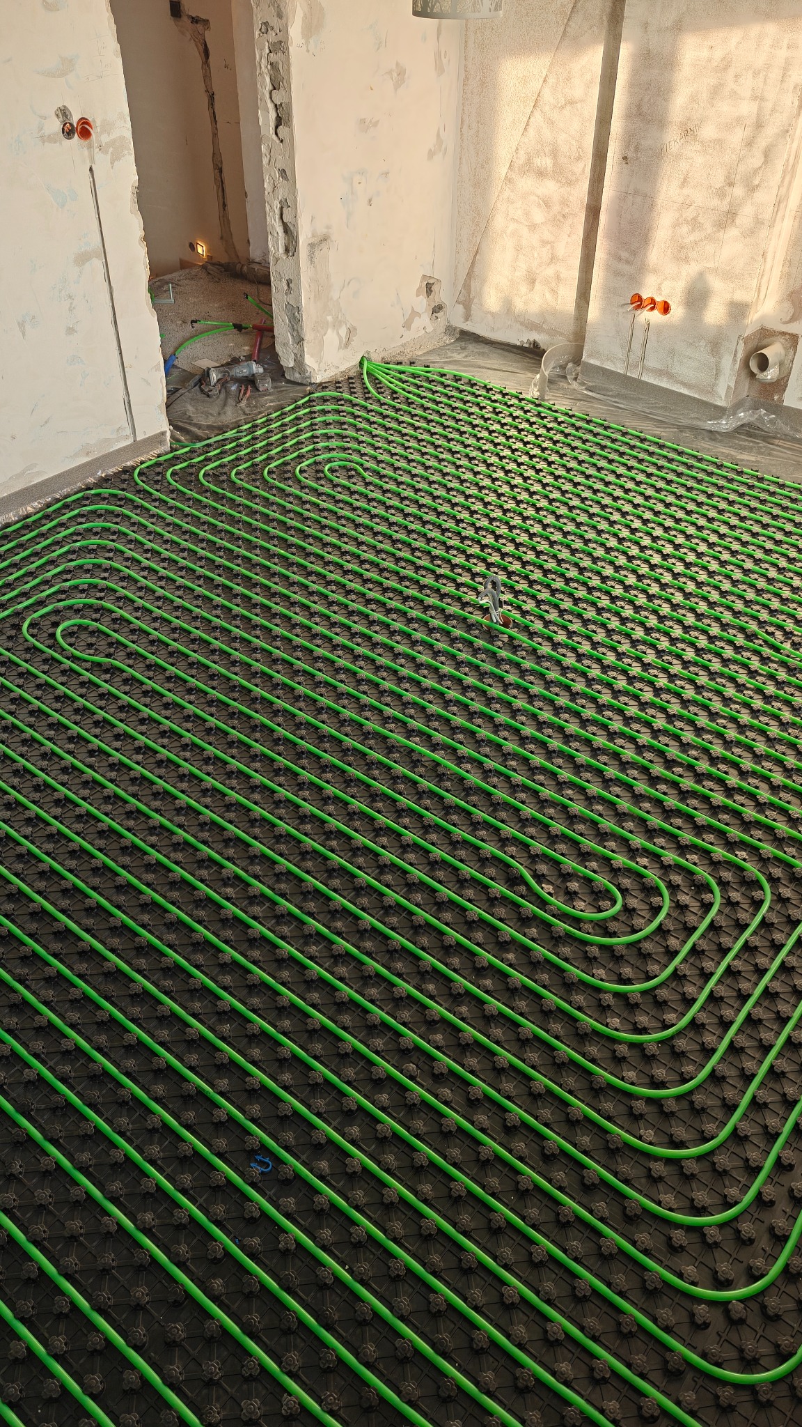 Zielone rurki ogrzewania podłogowego ułożone na czarnej macie w surowym wnętrzu. Widoczne narzędzia i instalacje elektryczne w ścianie. Ujęcie z góry, ukośne.