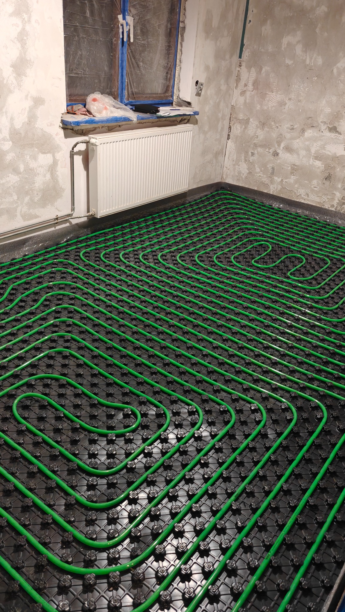 Zielone rury ogrzewania podłogowego ułożone na czarnej macie w surowym wnętrzu z grzejnikiem i oknem zabezpieczonym folią malarską. Widok z góry.