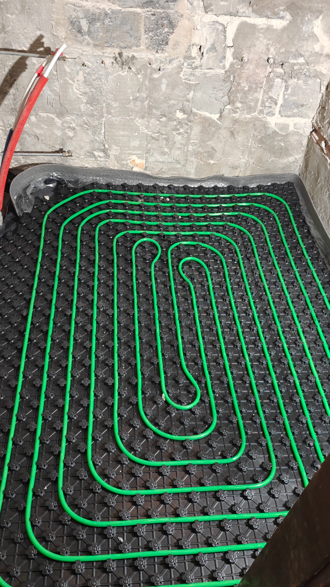 Ułożone, zielone rury ogrzewania podłogowego na czarnej macie systemowej, widoczne przyłącza do ściany w surowym stanie.