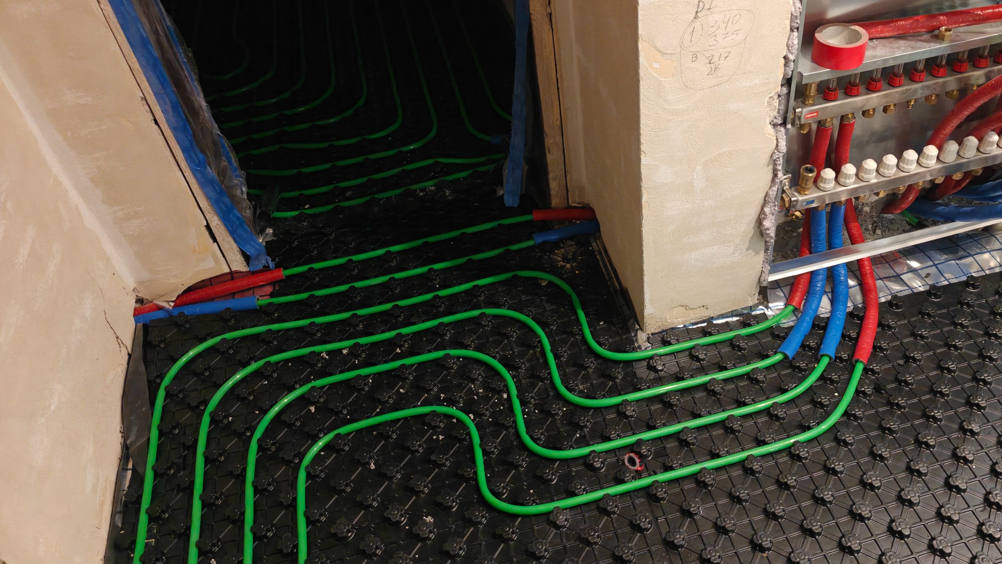 Ułożone, zielone rury ogrzewania podłogowego na czarnej macie montażowej, z widocznym rozdzielaczem i fragmentem ściany z notatkami. Instalacja przed zalaniem posadzką.