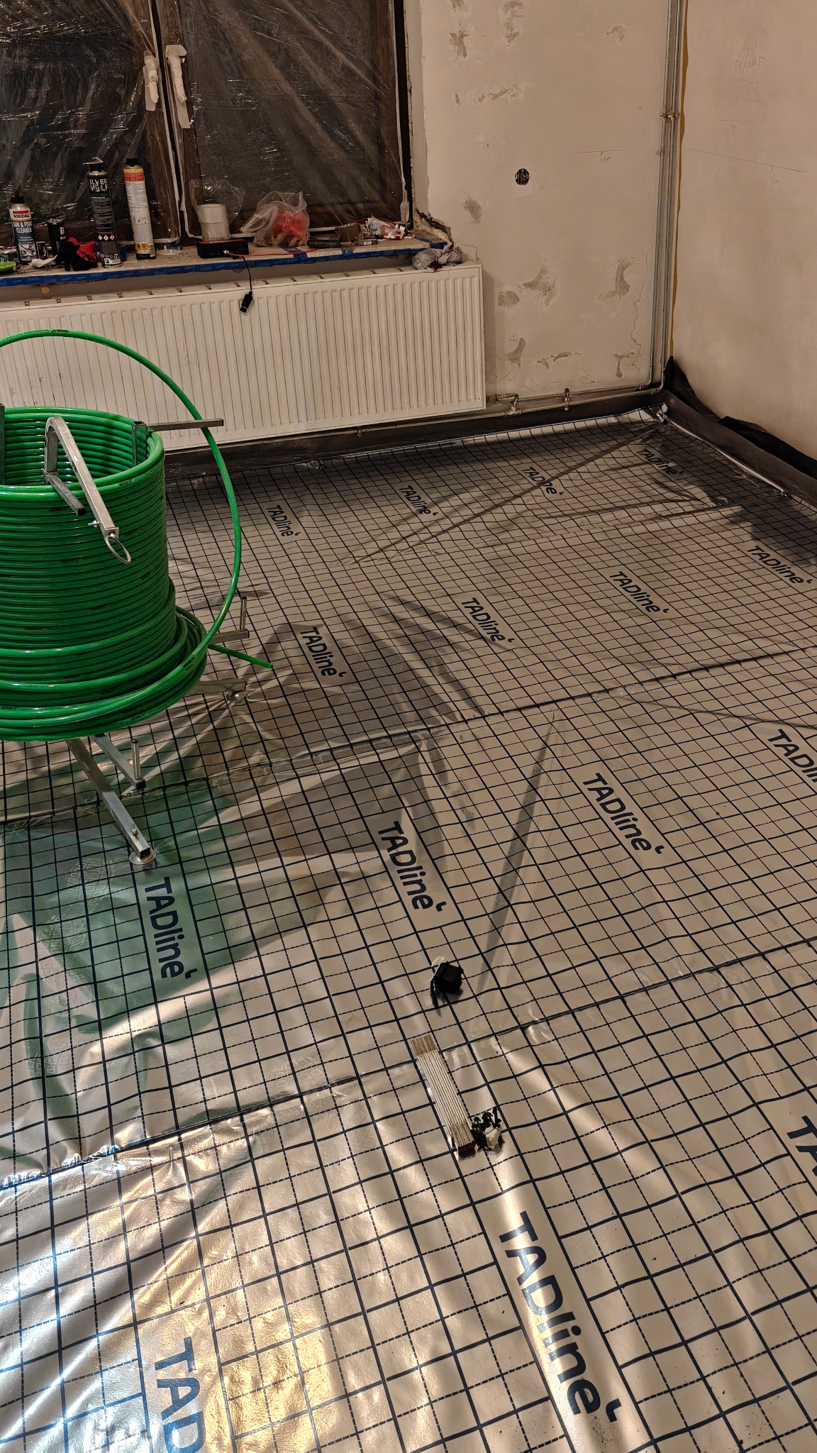 Rozłożona mata grzewcza TAdline z rolką zielonych rur na metalowym stojaku w pomieszczeniu w trakcie remontu. Widoczne elementy instalacji grzewczej.