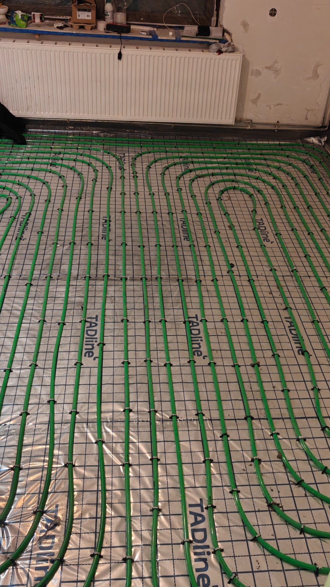 Ułożone, zielone rury ogrzewania podłogowego marki TAdline na folii z siatką, widoczne czarne uchwyty mocujące. W tle grzejnik i fragment ściany w trakcie remontu.