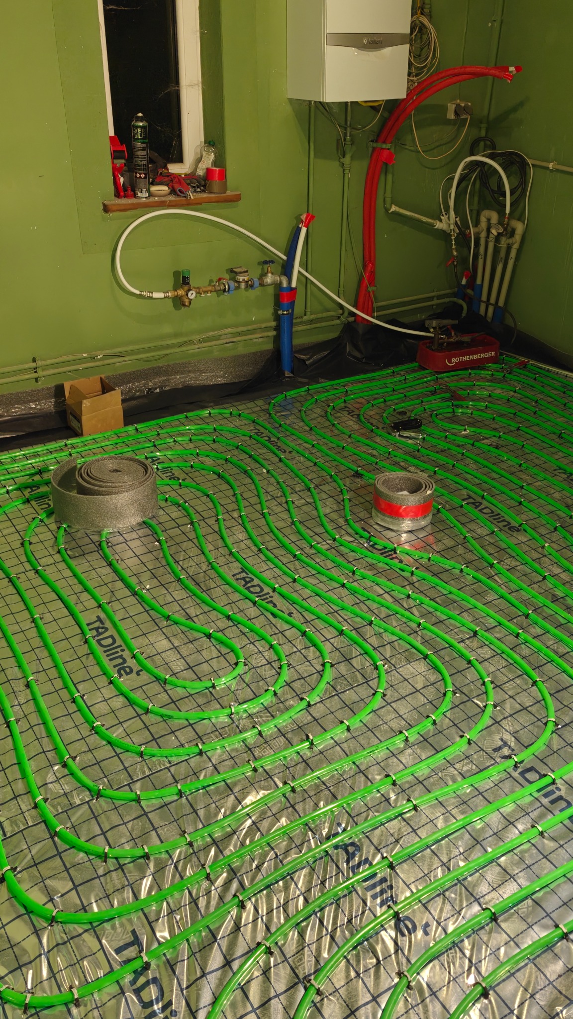 Zielone rury ogrzewania podłogowego ułożone na folii w Świętochłowicach. Widoczny rozdzielacz i narzędzia instalacyjne, okno z akcesoriami.