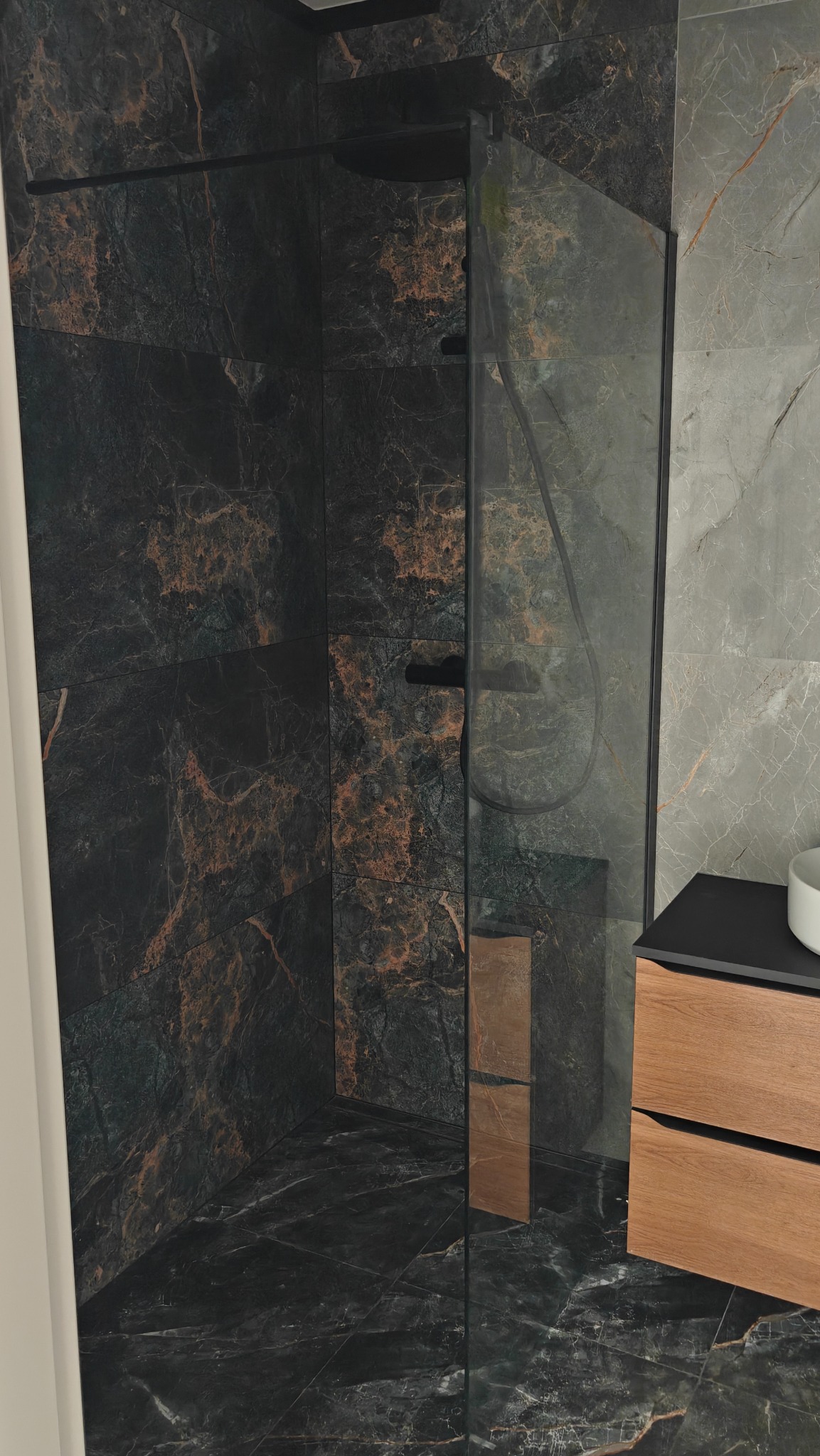 Nowoczesna kabina prysznicowa z czarnymi profilami i marmurowymi płytkami w odcieniach czerni i złota. Widoczna armatura i szafka łazienkowa z drewnianymi frontami.